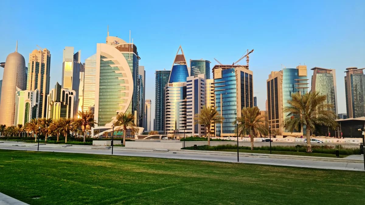 Glass skyscrapers of the West Bay skyline in Doha under a clear blue sky, with palm trees and a lawn in the foreground