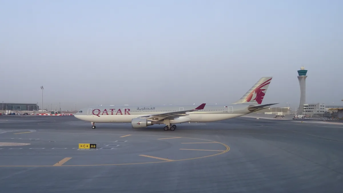 A Qatar Airways Airbus A330 in gray and burgundy livery taxiing across the apron at Hamad International Airport, with the airport control tower behind it