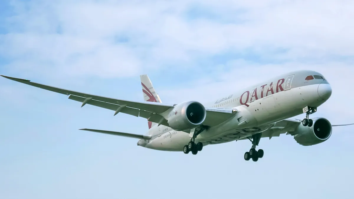 A Qatar Airways Boeing 787-8 in maroon and white livery flying with its landing gear down against a pale cloudy sky
