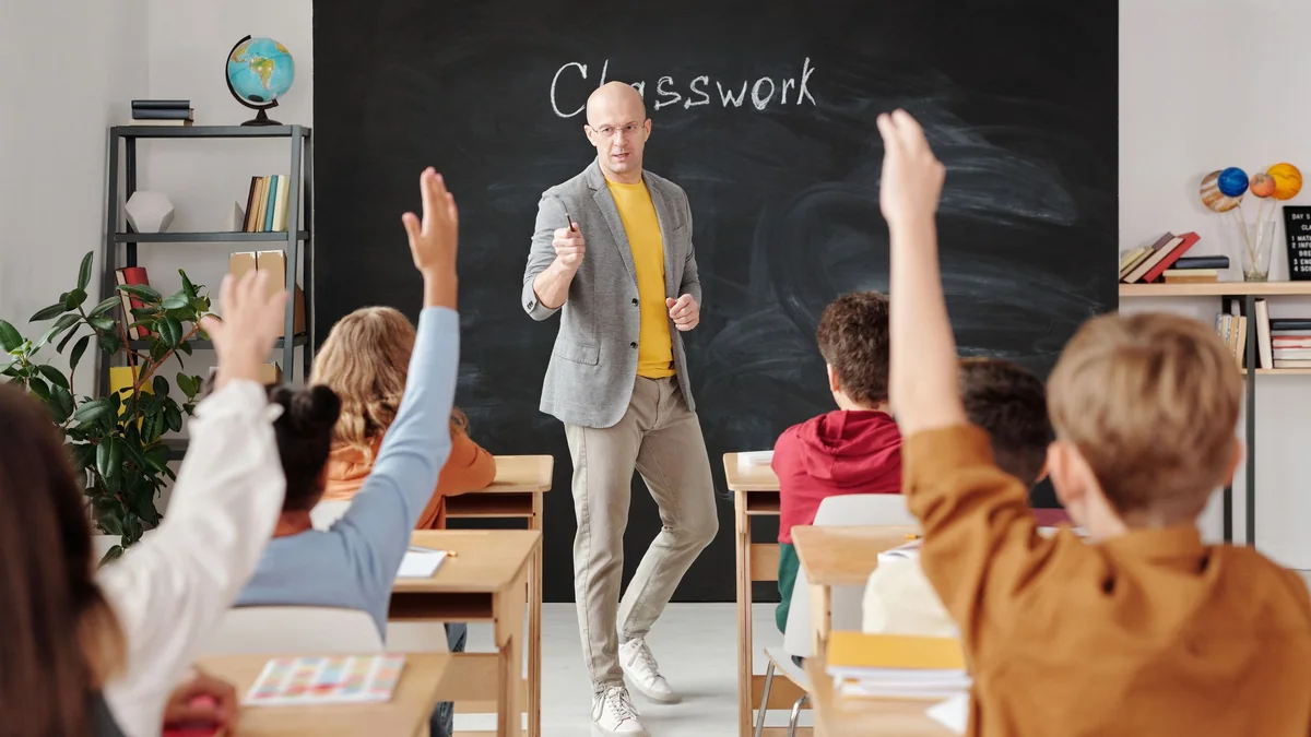 A teacher in a grey blazer stands in front of a chalkboard reading Classwork while students at wooden desks raise their hands