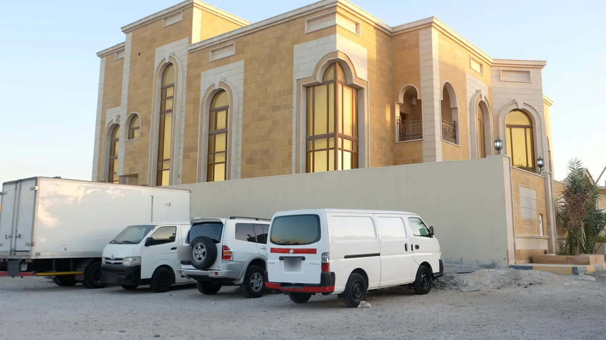 A white box truck, a minibus, an SUV and a white panel van parked beside a villa wall in Abu Hamour, Doha