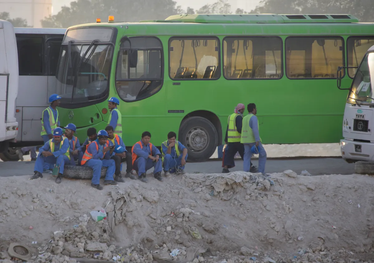 Foreign workers in blue overalls and hard hats resting beside buses at a construction site at The Pearl, Doha