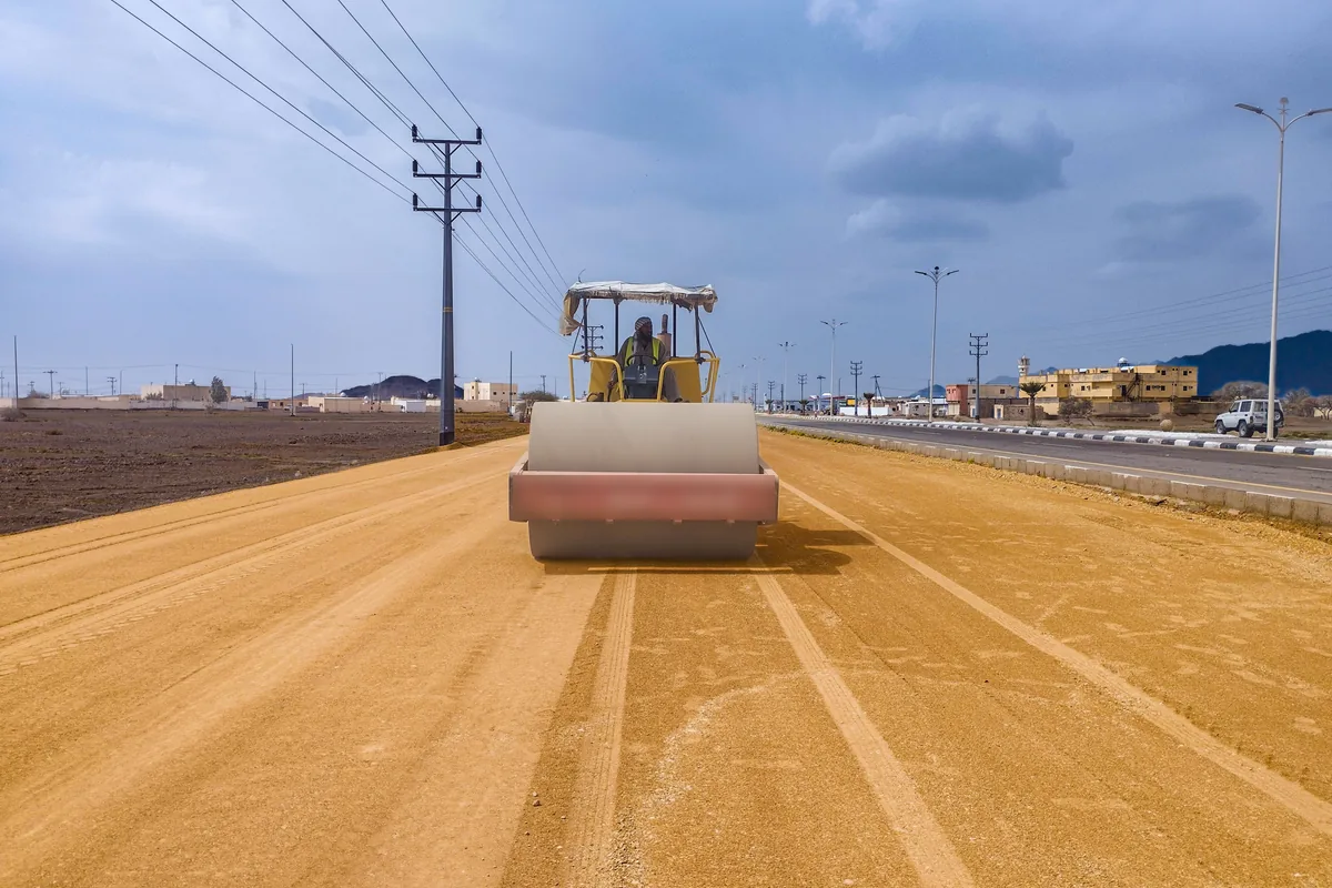 Road roller compacting the sandy base of a new road beside an existing highway, with power lines and low buildings under a cloudy sky
