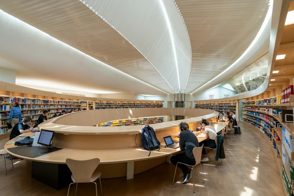 Students studying at laptops around an oval desk in a modern university library with curved bookshelves and a lit wooden ceiling
