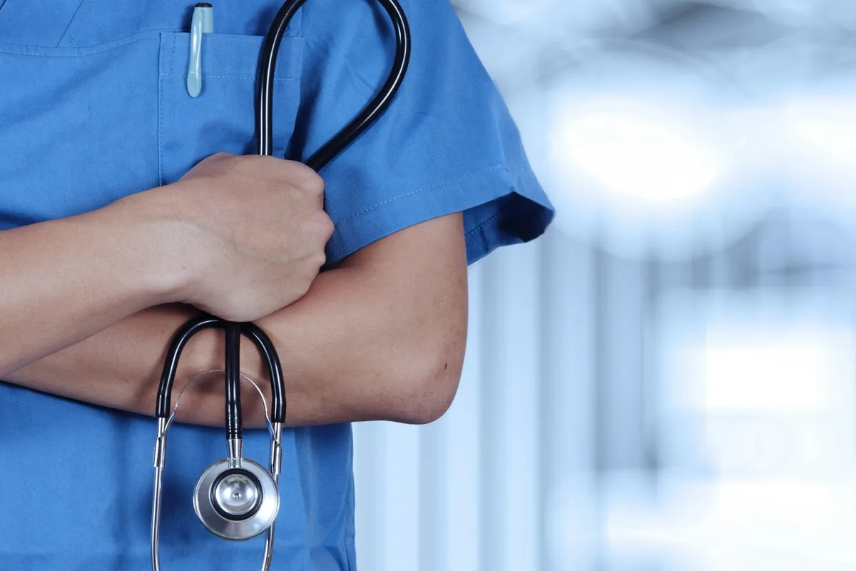 Healthcare worker in blue scrubs with arms crossed, holding a stethoscope against a blurred hospital background