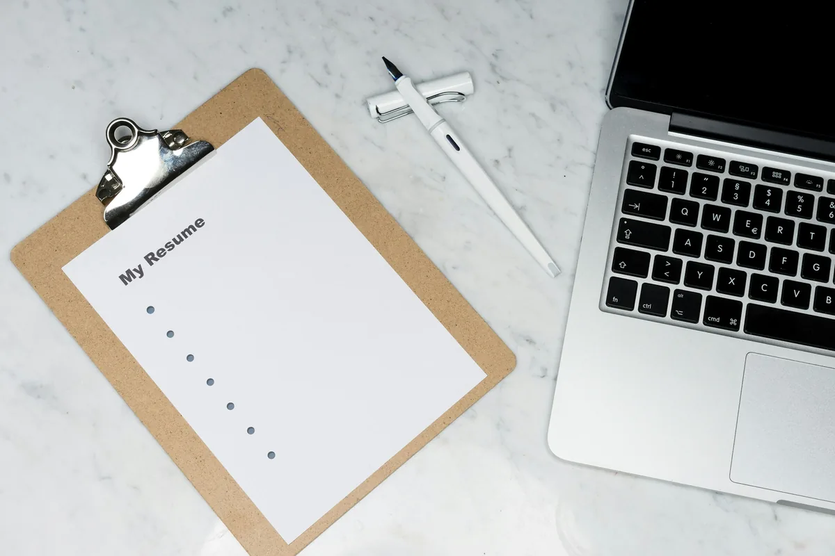 Clipboard holding a blank resume template next to a pen and an open laptop on a marble desk