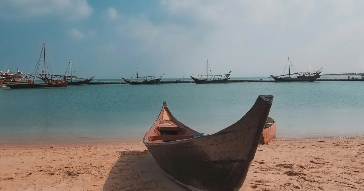 A traditional wooden boat on the sand at Katara Beach with dhows moored in the calm water behind