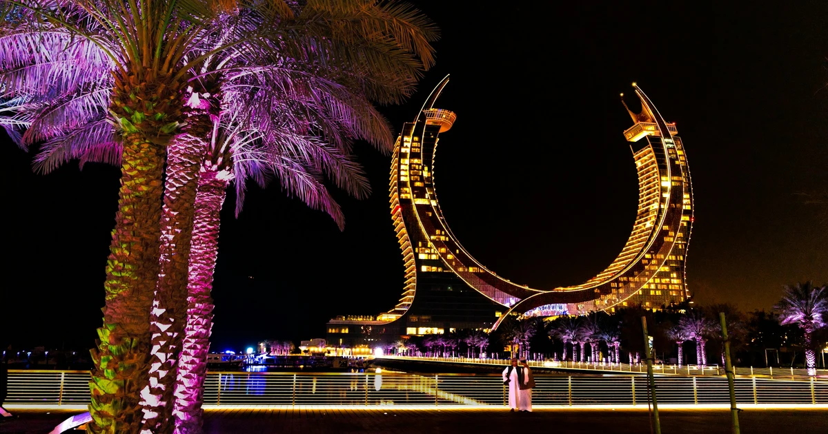 The crescent shaped Katara Towers lit up at night, seen from the Lusail waterfront promenade with illuminated palm trees