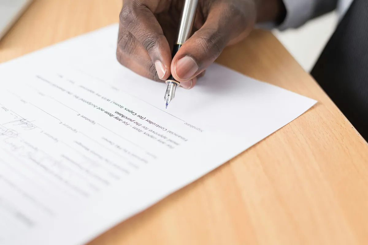 Hand holding a fountain pen over a printed document with signature lines on a wooden desk