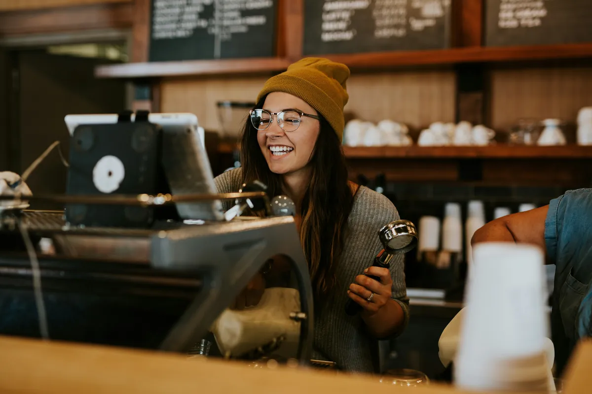 Smiling barista in glasses and a mustard beanie holding a portafilter behind a caf&eacute; counter