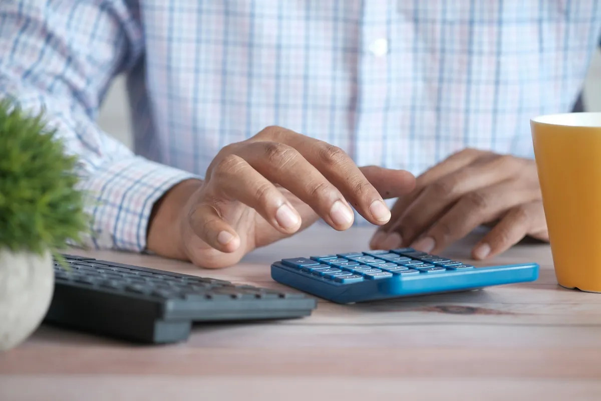 Hands of a person in a checked shirt using a blue calculator on a wooden desk beside a keyboard