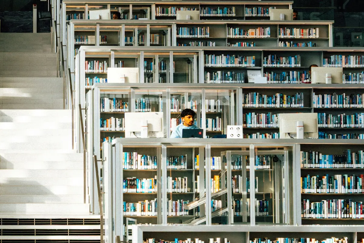 Young man studying on a laptop among tiered white bookshelves beside wide steps in a modern library