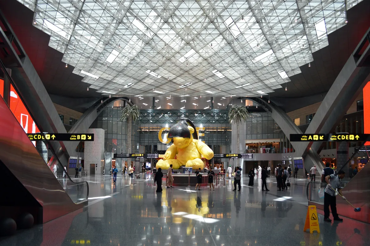 A large yellow teddy bear sculpture under a lamp in the duty-free hall at Hamad International Airport, with escalators, palm trees and gate signs around it