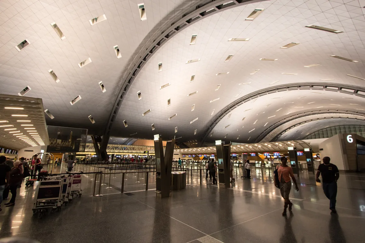 Passengers with luggage carts walk across the check-in hall at Hamad International Airport beneath a curved, vaulted ceiling, with check-in row 6 on the right