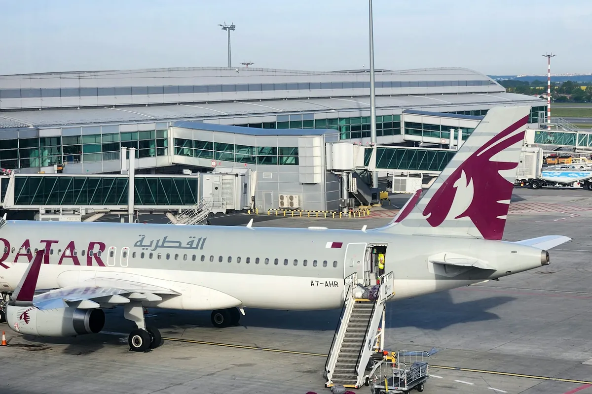 A Qatar Airways narrow-body aircraft parked at a stand with mobile boarding stairs at its front door, with jet bridges and a glass terminal behind
