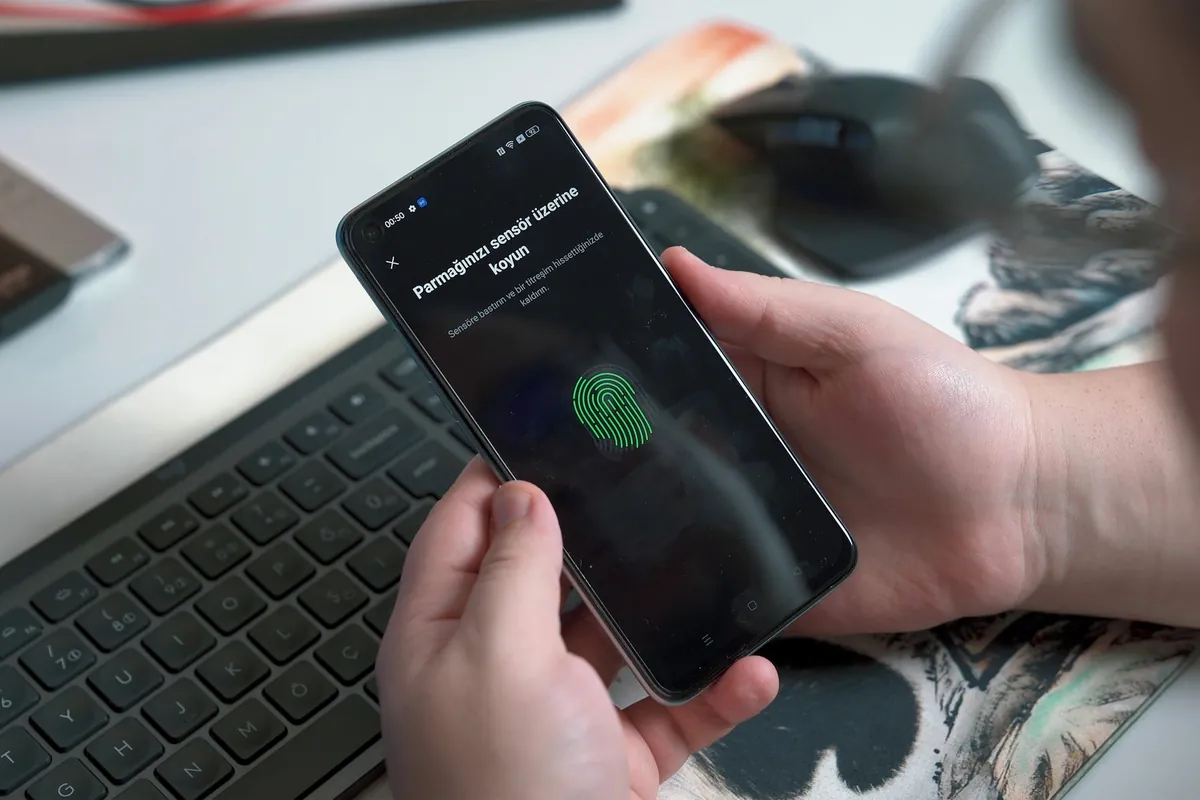 A hand holding a smartphone that shows a fingerprint verification prompt, above a laptop keyboard
