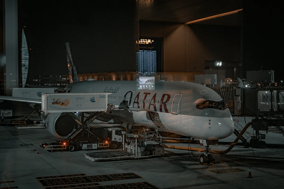 A Qatar Airways wide-body aircraft at a gate at night, connected to a jet bridge with a catering truck and ground equipment beside it