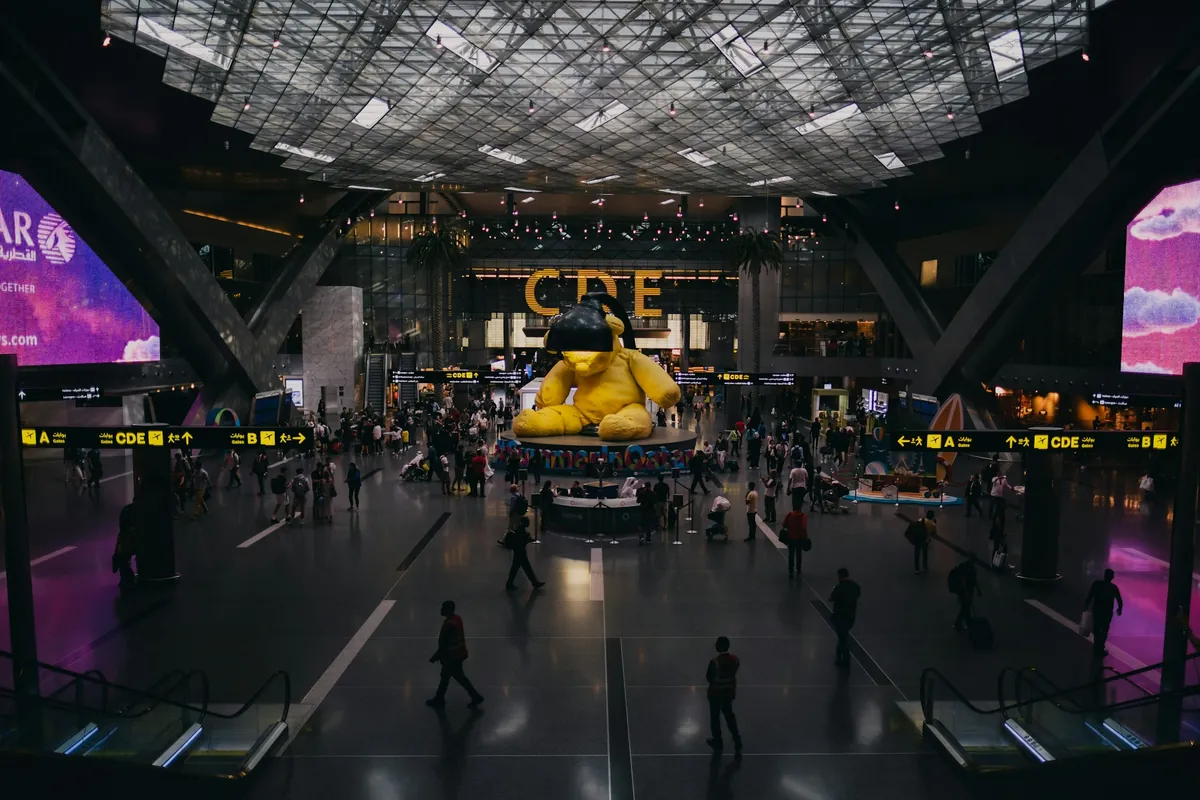 A large airport concourse with a lattice ceiling, a giant yellow teddy bear sculpture under a lamp, travelers walking and illuminated gate signs