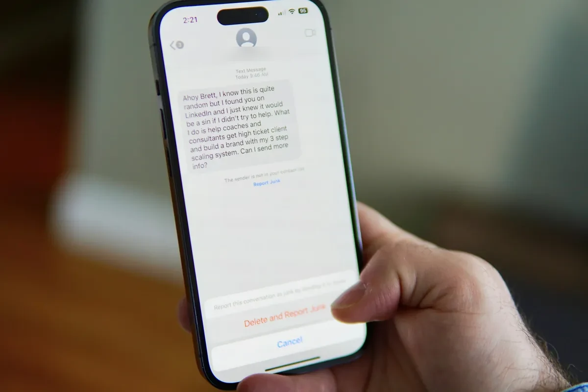 A hand holding a smartphone showing an unsolicited text message with a Delete and Report Junk option