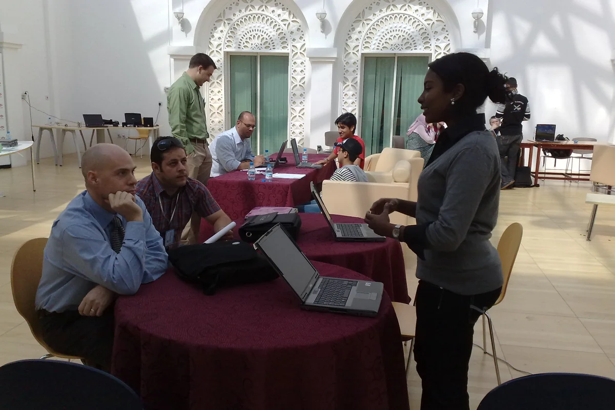 Adults with laptops seated at round tables listen to a woman speaking during an education conference workshop in Doha